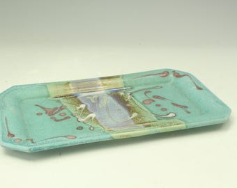 May include: A rectangular ceramic tray featuring a light turquoise glaze with a central band of blended blue, purple, and brown tones. The surface is decorated with scattered abstract splatters in dark pink and white, creating a textured appearance.
