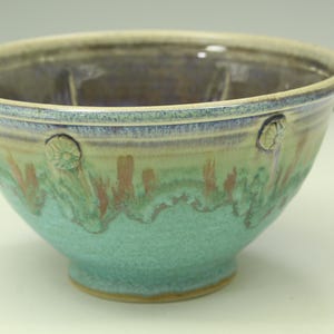 May include: A ceramic bowl featuring a wide rim and a footed base. The exterior displays a gradient of turquoise and light green with vertical brown streaks. Small circular floral-like embossed details are spaced evenly around the upper rim.