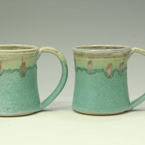 May include: Two ceramic mugs featuring a textured teal green base that transitions into a lighter cream-colored rim. Each mug has a curved handle and a decorative pattern of small, vertical, pinkish-brown brushstroke accents along the upper edge.