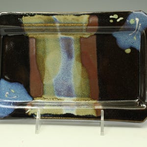 May include: A rectangular ceramic tray with a glossy black glaze featuring a central panel of blended tan, brown, and blue tones. The edges are accented with abstract blue and cream-colored brushwork patterns against the dark, reflective surface.