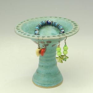May include: A light blue ceramic jewelry stand with a wide, perforated rim. A blue beaded bracelet rests inside the bowl, while two pairs of earrings, one with red and cream hearts and one with green star charms, hang from the rim.