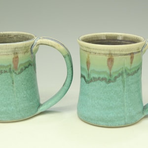 May include: Two handmade ceramic mugs with a turquoise glaze. The mugs have a cylindrical shape with a handle and a decorative band of green, brown, and cream colors near the rim. The mugs are suitable for hot or cold beverages.