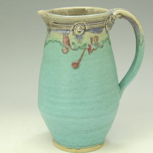 May include: Handmade ceramic pitcher in a soft turquoise glaze. The pitcher features a curved handle and a spout for easy pouring. Decorative elements include red and purple accents near the rim. A unique piece for serving beverages.