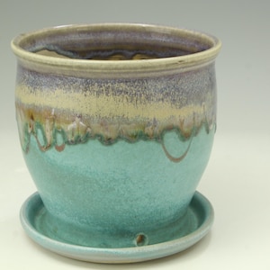 May include: A ceramic planter with a matching saucer, featuring a rounded body and a wide rim. The glaze transitions from a smooth turquoise at the base to a layered band of cream, brown, and muted purple tones near the top opening.