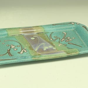 Puede incluir: Una bandeja rectangular de cer&aacute;mica con base turquesa y dise&ntilde;o decorativo. La bandeja presenta una secci&oacute;n central con acentos verdes, morados y marrones, y patrones florales blancos y marrones. Sus dimensiones son de unos 25 cm de largo y 13 cm de ancho.