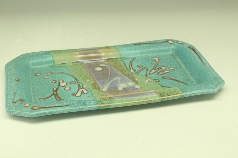 Puede incluir: Una bandeja rectangular de cer&aacute;mica con base turquesa y dise&ntilde;o decorativo. La bandeja presenta una secci&oacute;n central con acentos verdes, morados y marrones, y patrones florales blancos y marrones. Sus dimensiones son de unos 25 cm de largo y 13 cm de ancho.