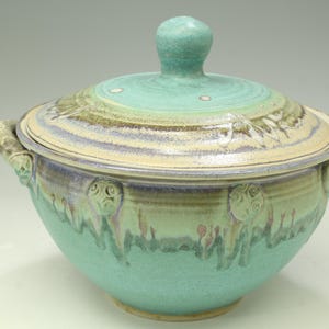 May include: A round ceramic lidded bowl featuring a turquoise and cream glaze with dripped blue and brown accents. The bowl has two side handles and a central knob on the lid. The surface displays a textured finish with circular embossed details.