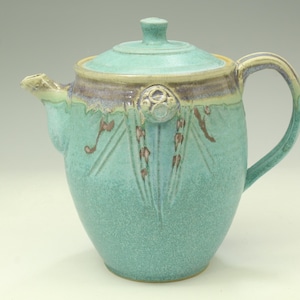 May include: A turquoise ceramic teapot with a matching lid, featuring a speckled glaze finish. The upper rim displays a band of muted purple and tan tones, with decorative raised circular and linear patterns extending down the body.