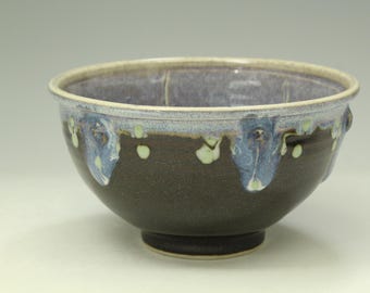 May include: A ceramic bowl with a dark brown base and a wide, rounded rim. The upper portion features a decorative glaze in shades of light blue and cream that drips down over the dark brown surface, creating a series of vertical patterns.