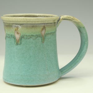 May include: A ceramic mug featuring a textured turquoise body with a smooth, light beige rim. The glaze drips down from the top edge, creating a layered effect. The mug has a wide, curved handle attached to the side and a flat, circular base.