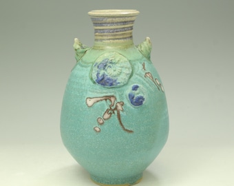 May include: A ceramic vase with a light turquoise glaze, featuring two small handles and a narrow neck with horizontal blue and white bands. The body is decorated with embossed circular patterns in blue and white, alongside brown linear markings.