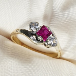 May include: A vintage ring with a gold band and a silver setting. The ring features a square, deep pink gemstone in the center, flanked by two smaller, clear gemstones. The ring is set against a cream-colored fabric.