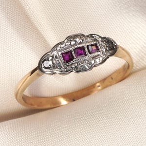 Puede incluir: Un anillo de oro con un engaste central alargado que contiene tres rubíes de color rosa intenso de corte cuadrado dispuestos en fila. El engaste es de metal plateado con detalles de milgrain y pequeños acentos decorativos en los bordes.