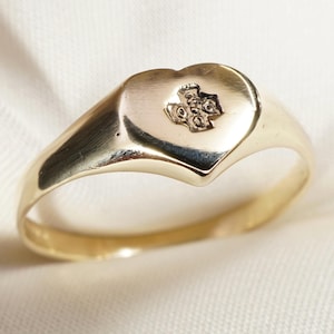 Peut inclure: Une bague en or avec une face en forme de cœur. La bague présente un petit emblème détaillé au centre du cœur. Le corps et la face de la bague ont une finition polie et réfléchissante. La bague est posée sur un fond blanc.