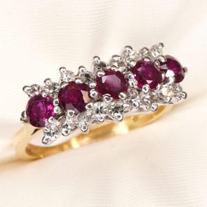 May include: A gold ring featuring a horizontal row of four round, deep red rubies set in white metal. The rubies are accented by small, clear, round-cut stones arranged in a cluster pattern surrounding each ruby, creating a decorative band design.