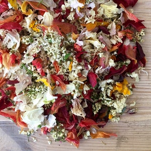 Pu&ograve; includere: Un mix di petali di fiori secchi in tonalit&agrave; di rosso, arancio, giallo, bianco e rosa. I petali sono sparsi su una superficie di legno.