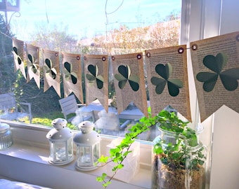 May include: A green shamrock banner made from paper pages hangs in a window. The banner is strung with twine and has metal eyelets. The shamrocks are cut out of the pages and are layered to create a 3D effect.
