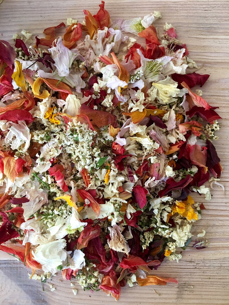 Pu&ograve; includere: Un mix di petali di fiori secchi in tonalit&agrave; di rosso, arancio, giallo, bianco e rosa. I petali sono sparsi su una superficie di legno.