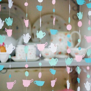 May include: A string of paper teacups and teapots in pink, blue, and white hangs in front of a shelf with teacups and saucers.