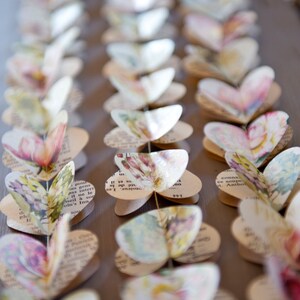 Puede incluir: Una guirnalda de mariposas de papel, cada una hecha de p&aacute;ginas de un libro antiguo, con patrones florales y texto. Las mariposas est&aacute;n enhebradas juntas con hilo y cuelgan en una guirnalda decorativa.