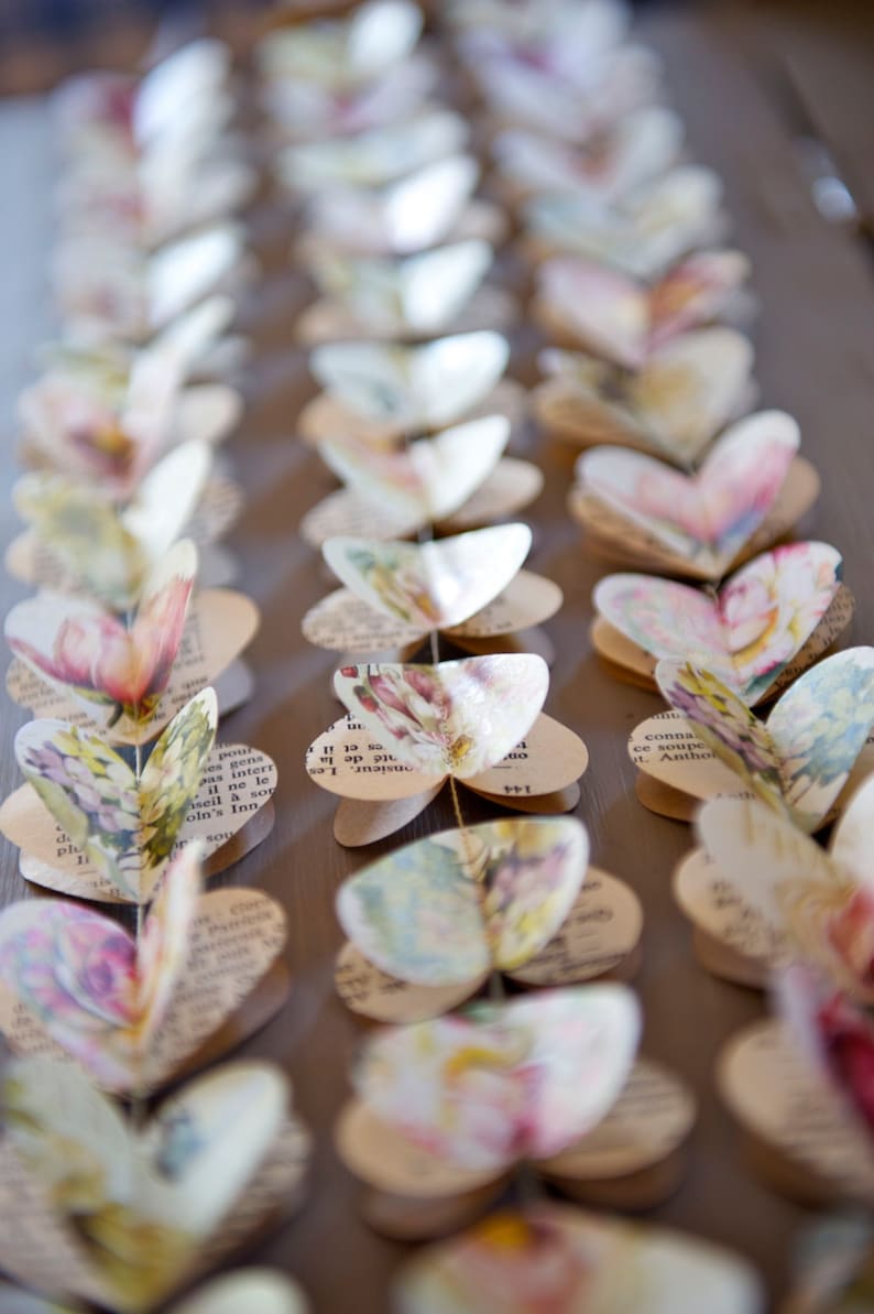 Puede incluir: Una guirnalda de mariposas de papel, cada una hecha de p&aacute;ginas de un libro antiguo, con patrones florales y texto. Las mariposas est&aacute;n enhebradas juntas con hilo y cuelgan en una guirnalda decorativa.