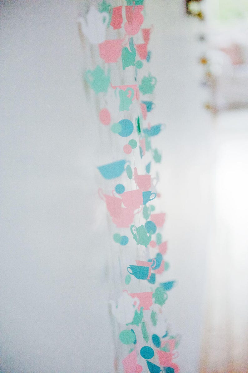 May include: A string of paper cutouts in the shape of teacups and other tea-related items. The cutouts are in shades of pink, blue, and green, and are strung together with white thread.