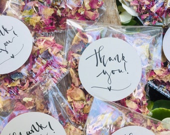 May include: Small white round stickers with the text "Thank you!" in black calligraphy on top of small clear bags filled with dried rose petals.