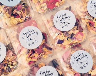 May include: A collection of small clear bags filled with colorful flower petals. Each bag has a round white sticker with black calligraphy that reads "Evelin & Johnny" and the date "05.07.19".