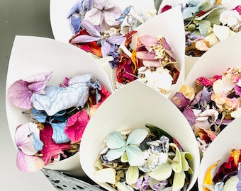 以下が含まれることがあります： ピンク、パープル、ブルー、グリーンの色合いの乾燥した花びらで満たされた白い紙の円錐形。円錐形は、装飾的なカットアウトデザインの金属製の容器に配置されています。