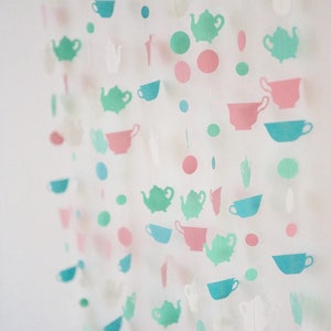 May include: A garland of paper cutouts featuring teacups and teapots in shades of blue, green, and pink. The cutouts are strung together with white thread and are hanging against a white background.