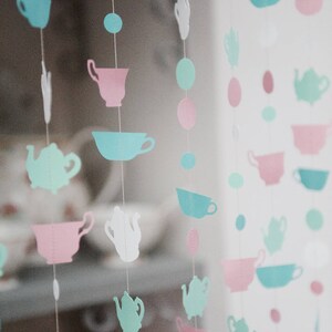 May include: A string of paper cutouts in the shape of teacups and teapots in shades of pink, blue, and green. The cutouts are strung on white thread and hang from a window.