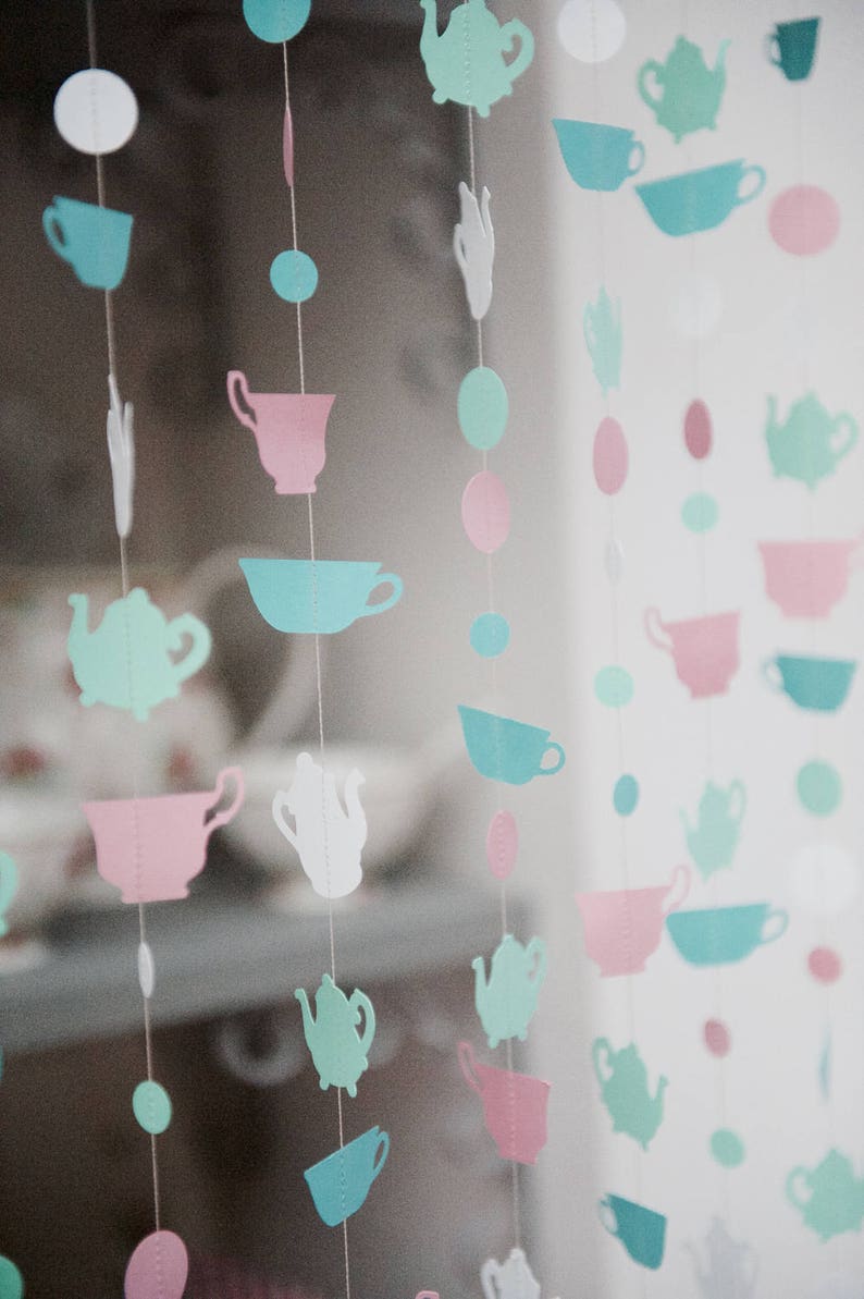 May include: A string of paper cutouts in the shape of teacups and teapots in shades of pink, blue, and green. The cutouts are strung on white thread and hang from a window.