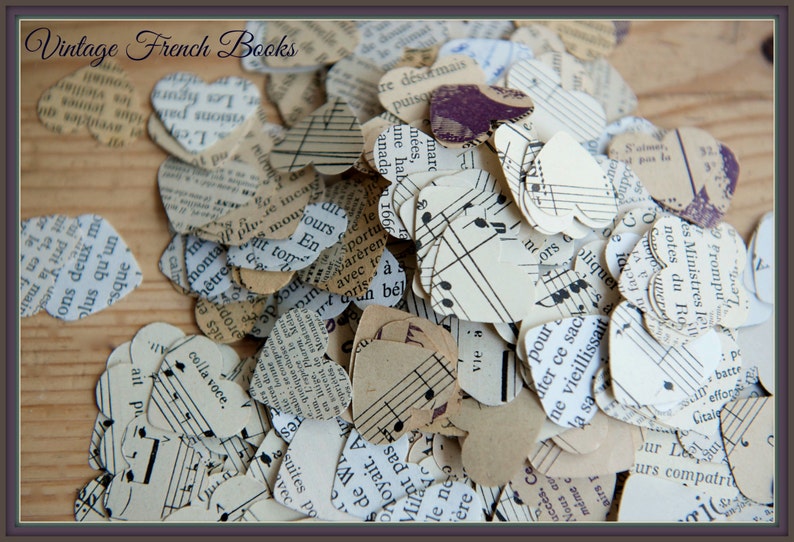 Puede incluir: Un mont&oacute;n de confeti en forma de coraz&oacute;n hecho de p&aacute;ginas de libros franceses antiguos y partituras musicales. El confeti est&aacute; esparcido sobre una superficie de madera. El texto "Vintage French Books" es visible en la parte superior de la imagen.