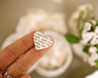 May include: A small, heart-shaped paper cutout with text printed on it is held between two fingers. The paper heart is white with black text and is set against a blurred background of white flowers and a neutral surface.