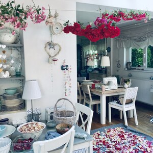 Pu&ograve; includere: Un tavolo bianco con una tovaglia bianca e un cesto di fiori. Ci sono petali di rosa rosa e rossi sparsi sul pavimento. Il tavolo &egrave; apparecchiato con un piatto bianco, un bicchiere e un tovagliolo. C'&egrave; una lampada bianca sul tavolo.