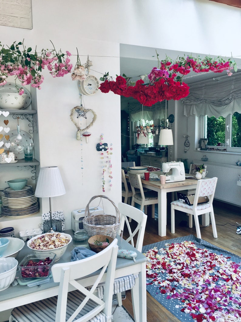 Pu&ograve; includere: Un tavolo bianco con una tovaglia bianca e un cesto di fiori. Ci sono petali di rosa rosa e rossi sparsi sul pavimento. Il tavolo &egrave; apparecchiato con un piatto bianco, un bicchiere e un tovagliolo. C'&egrave; una lampada bianca sul tavolo.