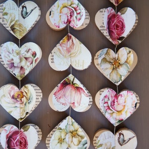 Puede incluir: Una guirlanda de corazones de papel con patrones florales y texto impreso en ellos. Los corazones est&aacute;n enhebrados juntos con un hilo dorado. La guirlande est&aacute; colgada en una superficie de madera.