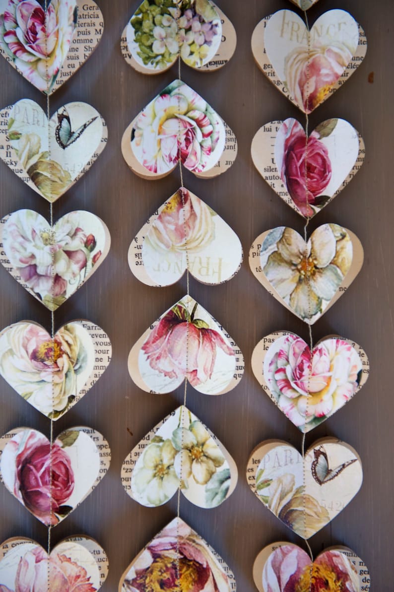 Puede incluir: Una guirlanda de corazones de papel con patrones florales y texto impreso en ellos. Los corazones est&aacute;n enhebrados juntos con un hilo dorado. La guirlande est&aacute; colgada en una superficie de madera.