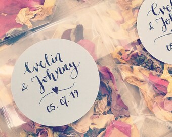 May include: Close-up of a small, clear plastic bag filled with dried flower petals. A white round sticker with black calligraphy reads "Evelyn & Johnny 05.07.19".