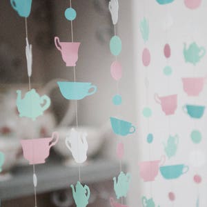 May include: A string of paper cutouts in the shape of teacups and teapots in shades of pink, blue, and green. The cutouts are strung on white thread and hang from a ceiling or wall.