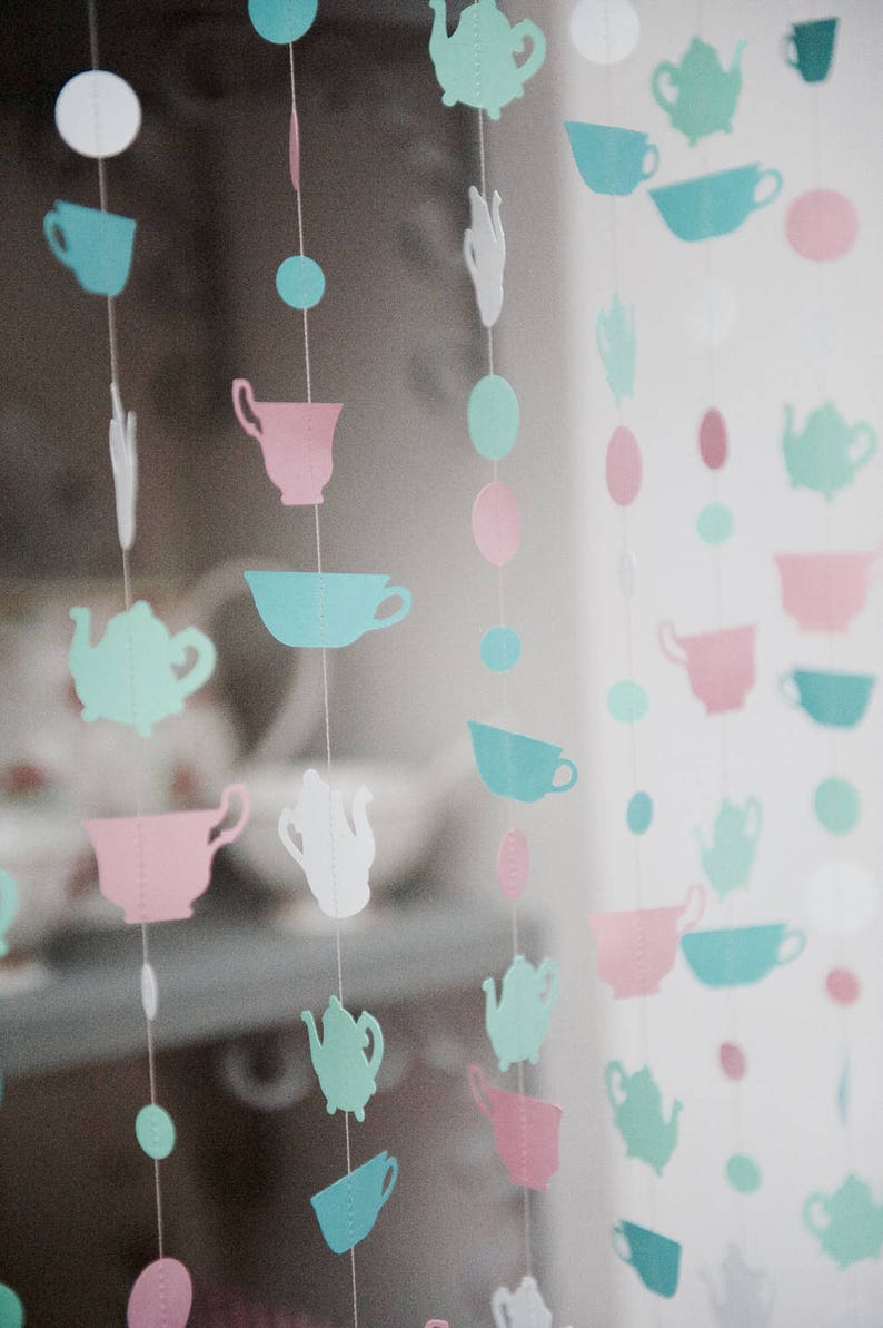 May include: A string of paper cutouts in the shape of teacups and teapots in shades of pink, blue, and green. The cutouts are strung on white thread and hang from a ceiling or wall.