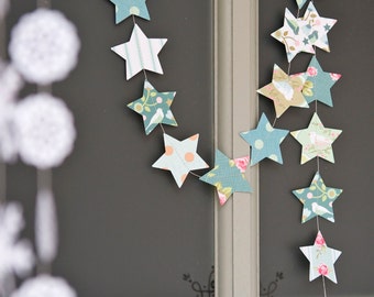 May include: A string of paper stars in various patterns and colours hangs on a window. The stars are connected by a thin string and are decorated with floral and geometric designs.