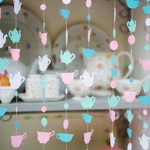 Puede incluir: Una guirnalda de tazas y teteras de papel en rosa, azul y verde colgando del techo. Las tazas y las teteras están recortadas en papel y enhebradas con hilo blanco.
