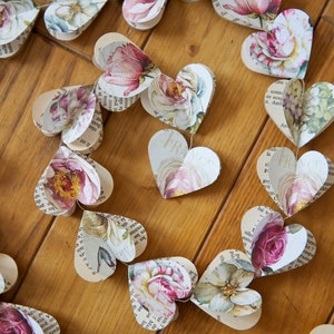 Puede incluir: Una guirnalda de corazones de papel con patrones florales y texto impreso en ellos. Los corazones est&aacute;n unidos con un cordel y cuelgan de una superficie de madera.