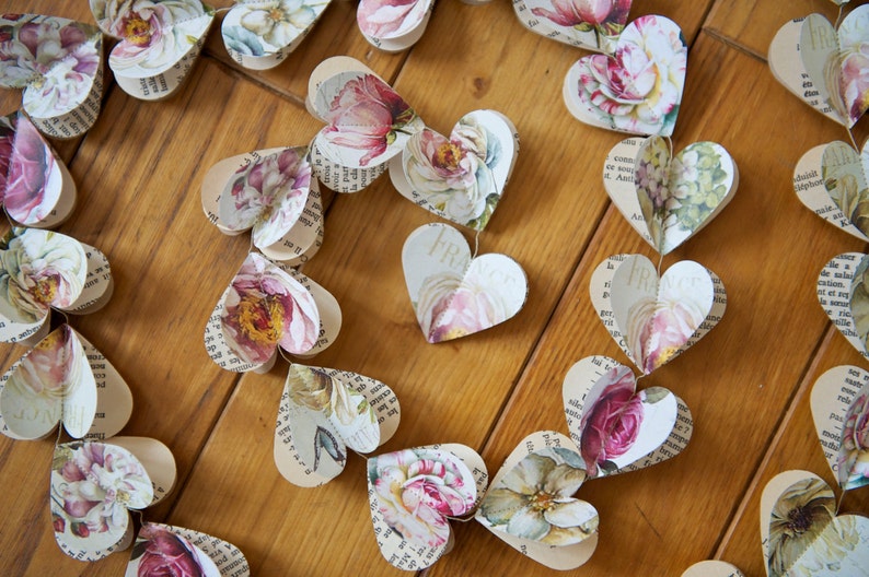 Puede incluir: Una guirnalda de corazones de papel con patrones florales y texto impreso en ellos. Los corazones est&aacute;n unidos con un cordel y cuelgan de una superficie de madera.