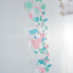 May include: A string of paper cutouts in the shape of teacups and teapots, alternating in pink, blue, and green colors. The string is hanging from the ceiling.