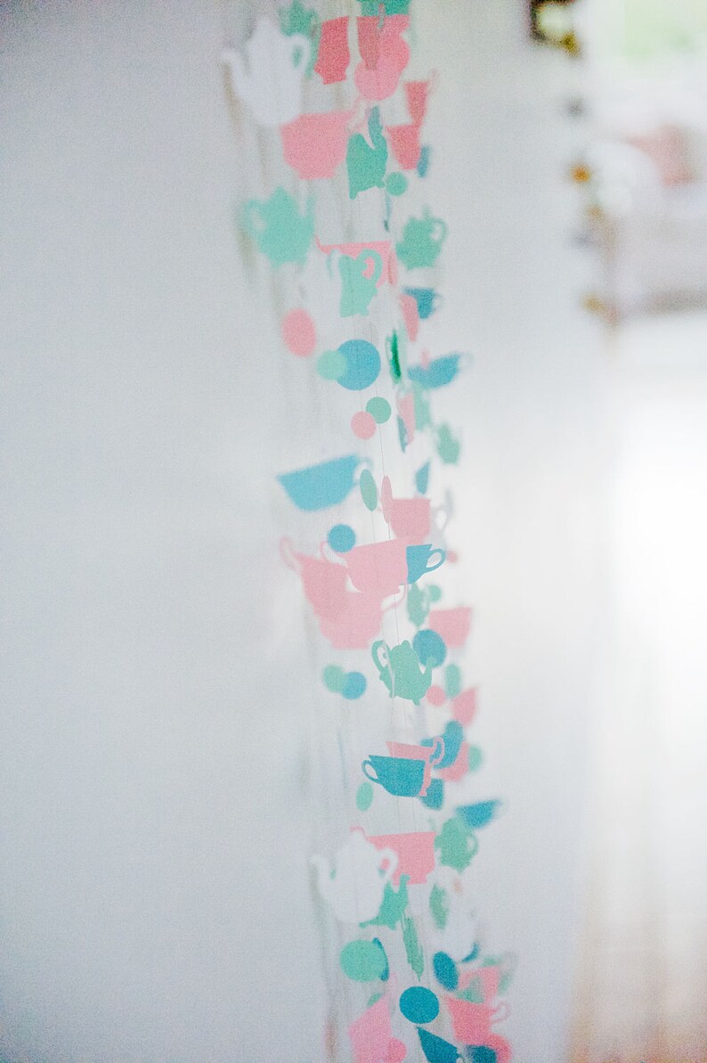 May include: A string of paper cutouts in the shape of teacups and teapots, alternating in pink, blue, and green colors. The string is hanging from the ceiling.