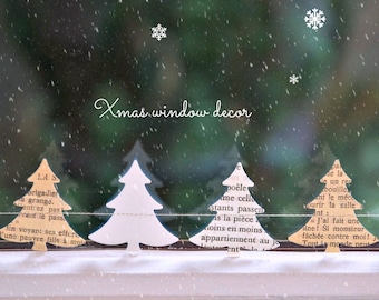 Può includere: Decorazione natalizia per finestre con alberi di Natale di carta in varie tonalità di bianco e beige, disposti in fila. Gli alberi sono ambientati su uno sfondo verde sfocato con fiocchi di neve che cadono. Il testo "Xmas window decor" è scritto in corsivo.