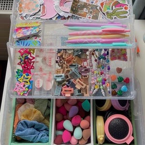 Può includere: Un organizer trasparente a più scomparti pieno di vari oggetti. La sezione superiore contiene adesivi e penne. La sezione centrale contiene fermagli per capelli e perline. La sezione inferiore ha spugne per il trucco e accessori per capelli.