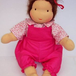 Puede incluir: Una muñeca hecha a mano con cabello castaño que lleva un atuendo rosa y blanco. La muñeca tiene una camisa rosa y blanca con lunares y un mono rosa. La muñeca está sentada sobre una superficie blanca.
